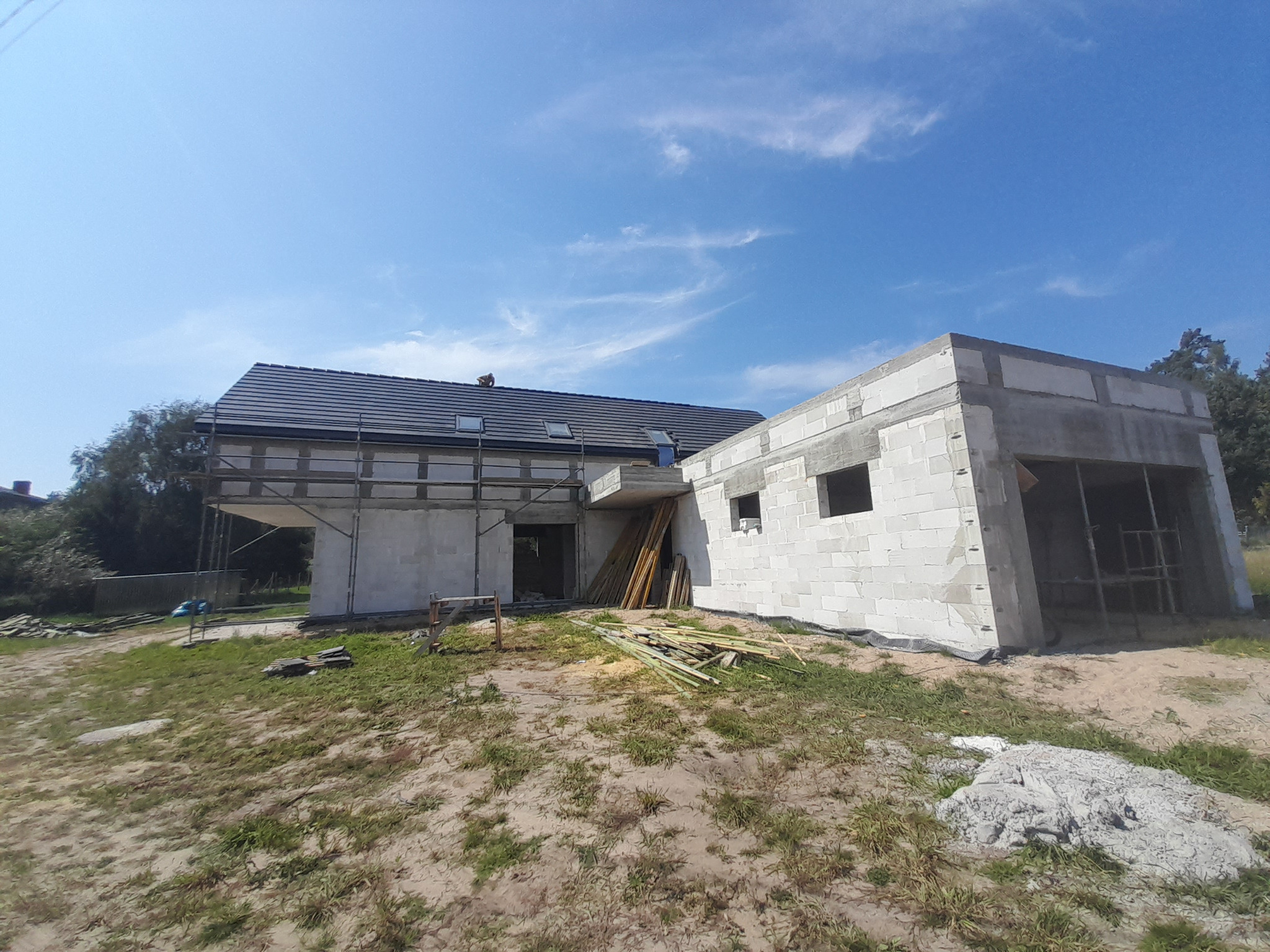 Surowy stan domu z białych bloczków, widoczne rusztowania i drewniane elementy konstrukcyjne, dach pokryty ciemną dachówką, błękitne niebo w tle.