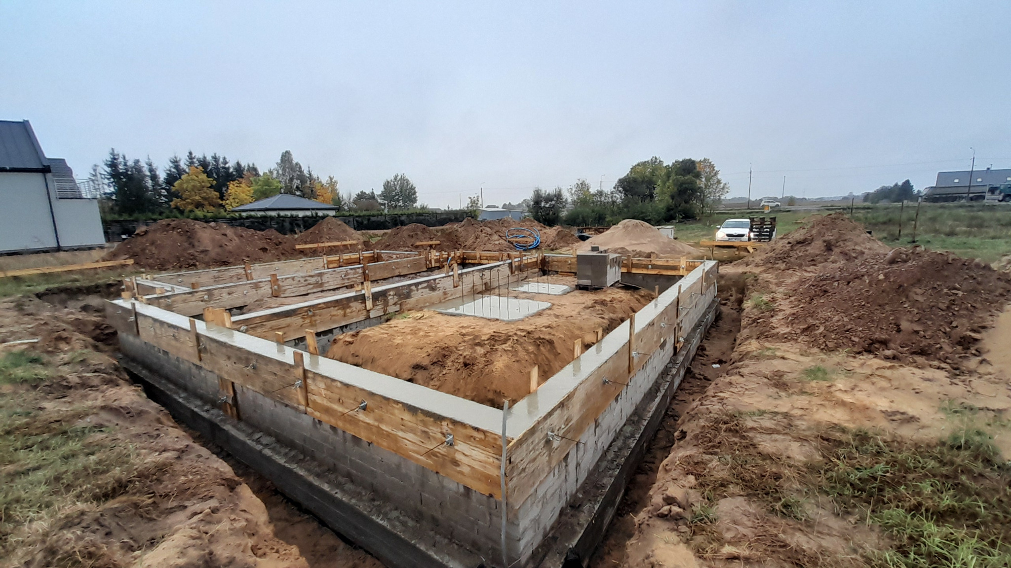 Wykopane fundamenty pod budynek z częściowo wylaną betonową wylewką, drewnianym szalunkiem i wystającymi prętami zbrojeniowymi, otoczone ziemią i widoczne z perspektywy szerokokątnej.