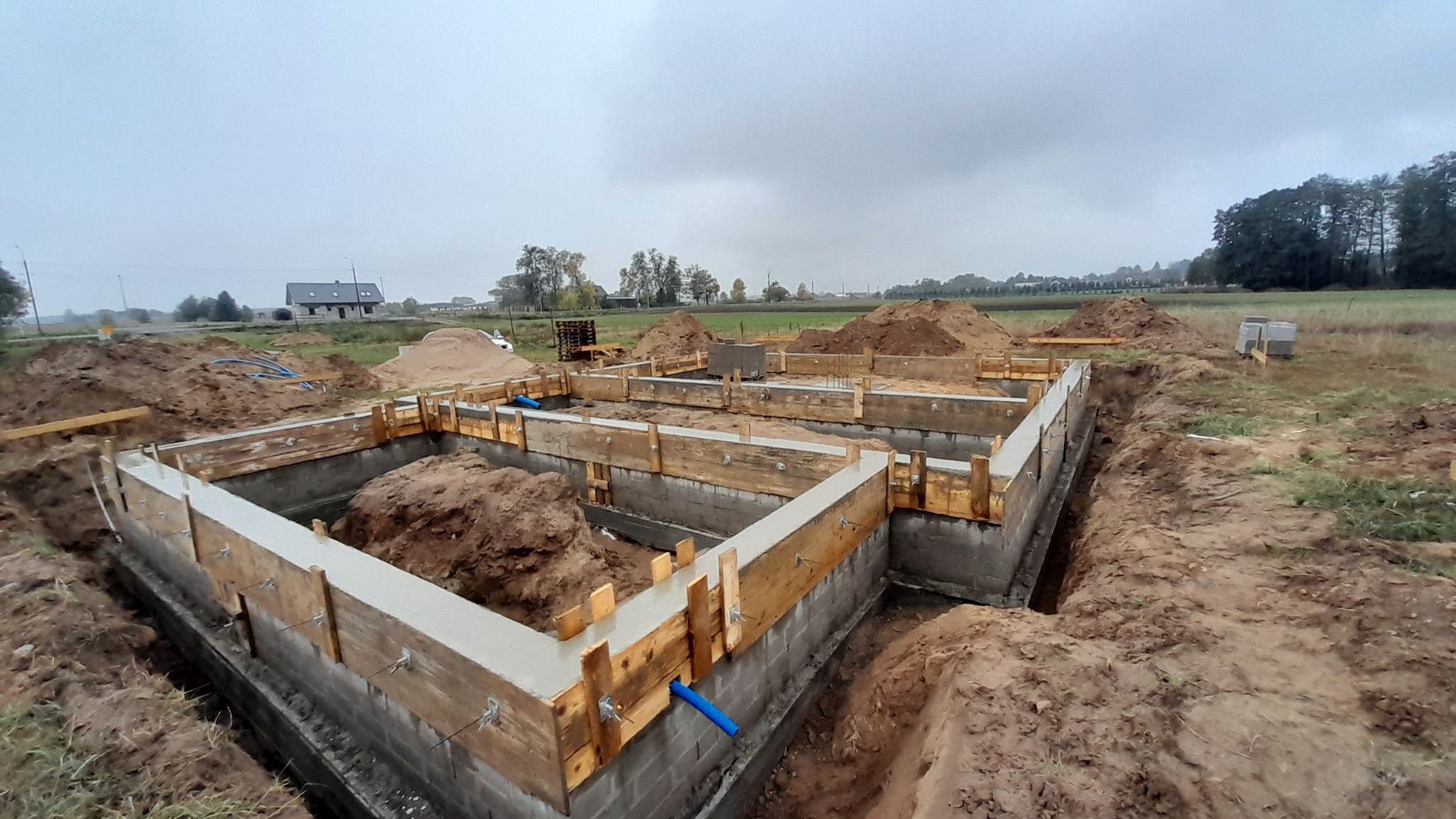 Wylane fundamenty pod budynek jednorodzinny, widoczne szalunki drewniane, ziemia wokół wykopu, na horyzoncie zabudowania.