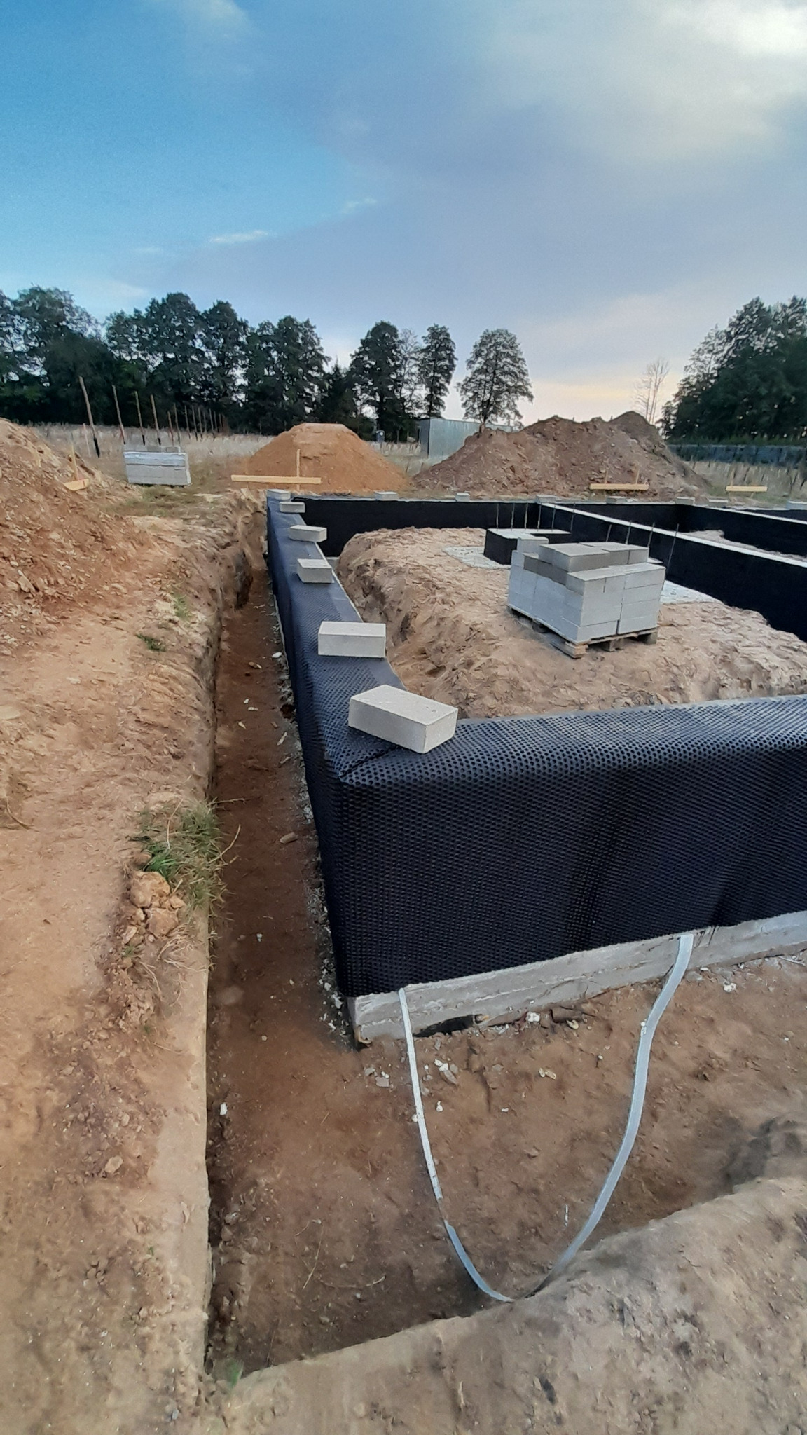 Fundamenty domu jednorodzinnego zabezpieczone folią kubełkową, widoczne wykopane rowy, przygotowane bloczki betonowe i pryzmy ziemi w tle.