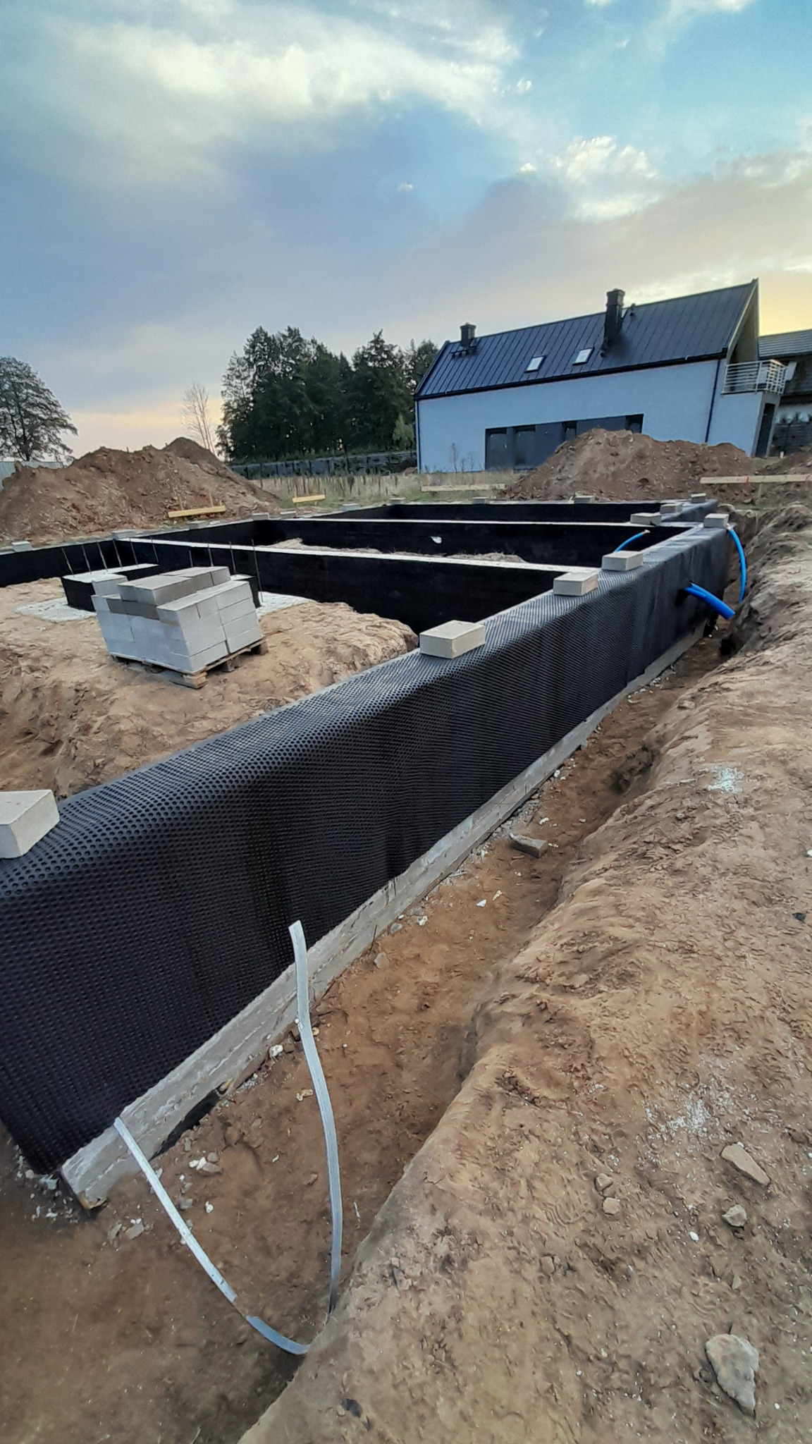 Wykop pod fundamenty z izolacją pionową z folii kubełkowej, widoczne bloczki betonowe i rury instalacyjne, w tle dom w trakcie budowy.