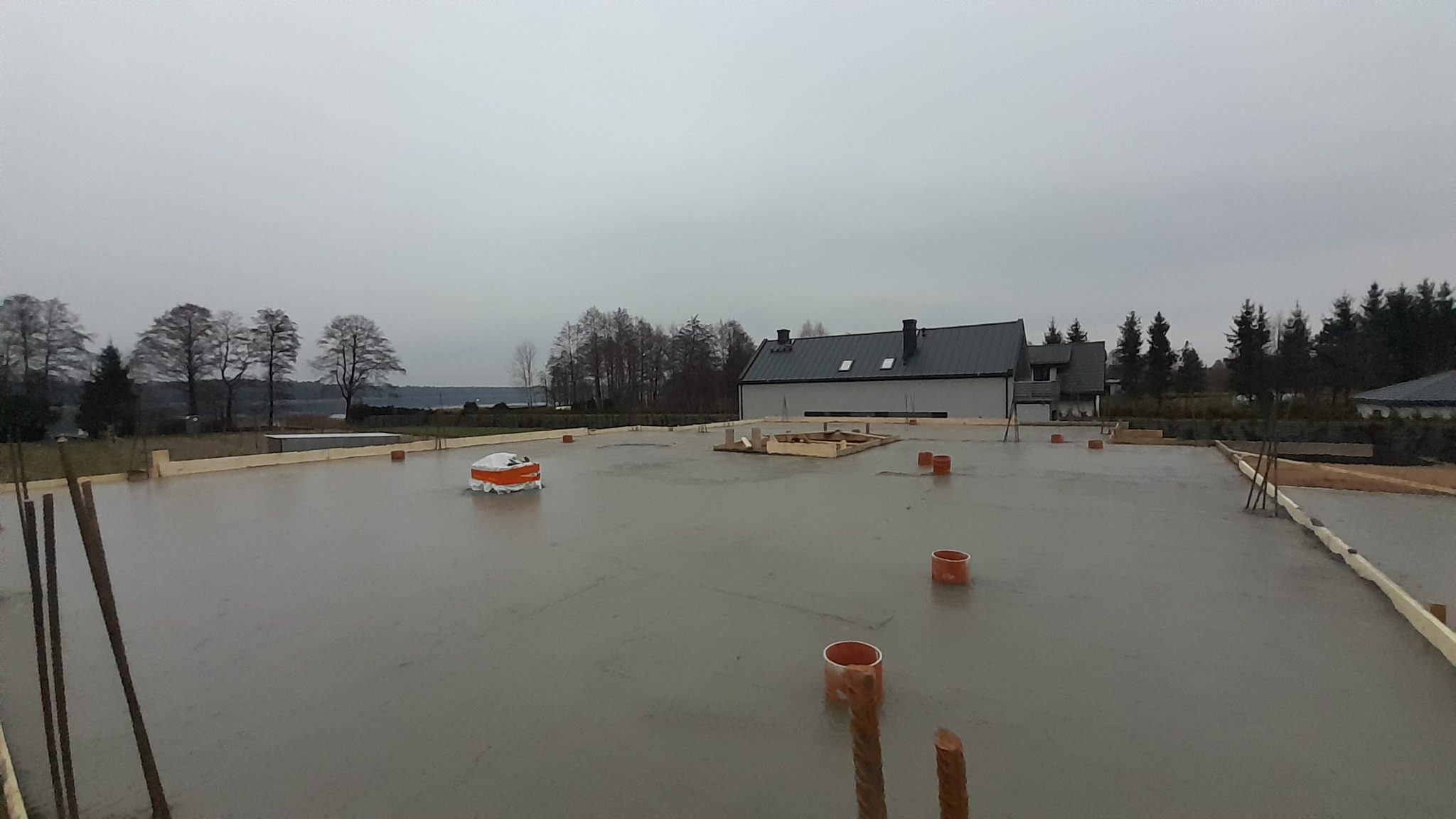 Wylany fundament pod dom jednorodzinny z widocznymi prętami zbrojeniowymi, deskowaniem i elementami instalacji, na tle zabudowy jednorodzinnej i pochmurnego nieba.