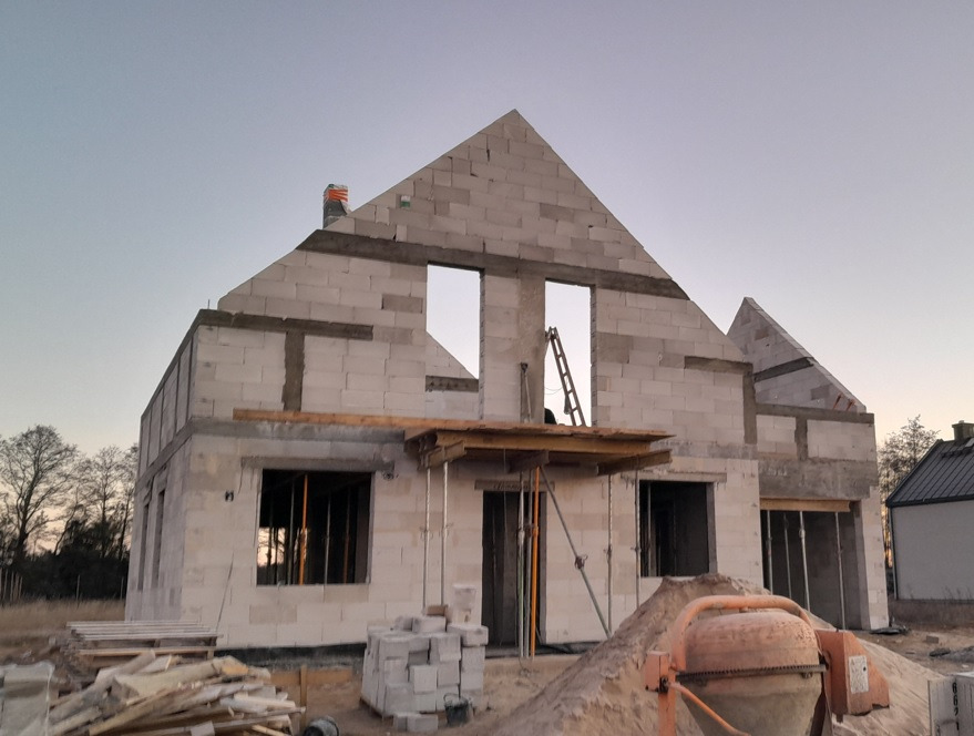 Surowy stan domu z białych bloczków podczas budowy, widoczne otwory okienne, drabina oparta o ścianę, sterta bloczków i betoniarka na pierwszym planie, niebo o zmierzchu w tle.