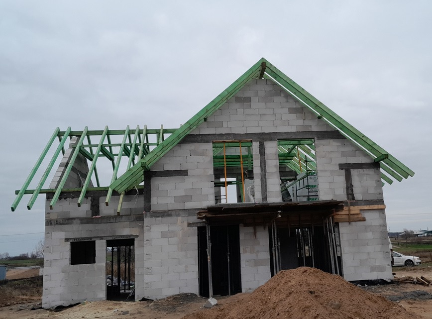 Surowy stan domu z betonu komórkowego w trakcie budowy, widoczne zielone elementy więźby dachowej i sterta piasku przed budynkiem.