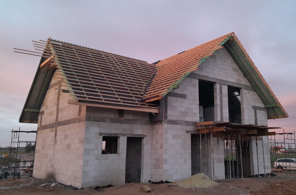 Niedokończony dom z białych bloczków, w trakcie budowy dachu z drewnianą konstrukcją i częściowo położoną dachówką, widoczne rusztowania i składowane materiały budowlane.