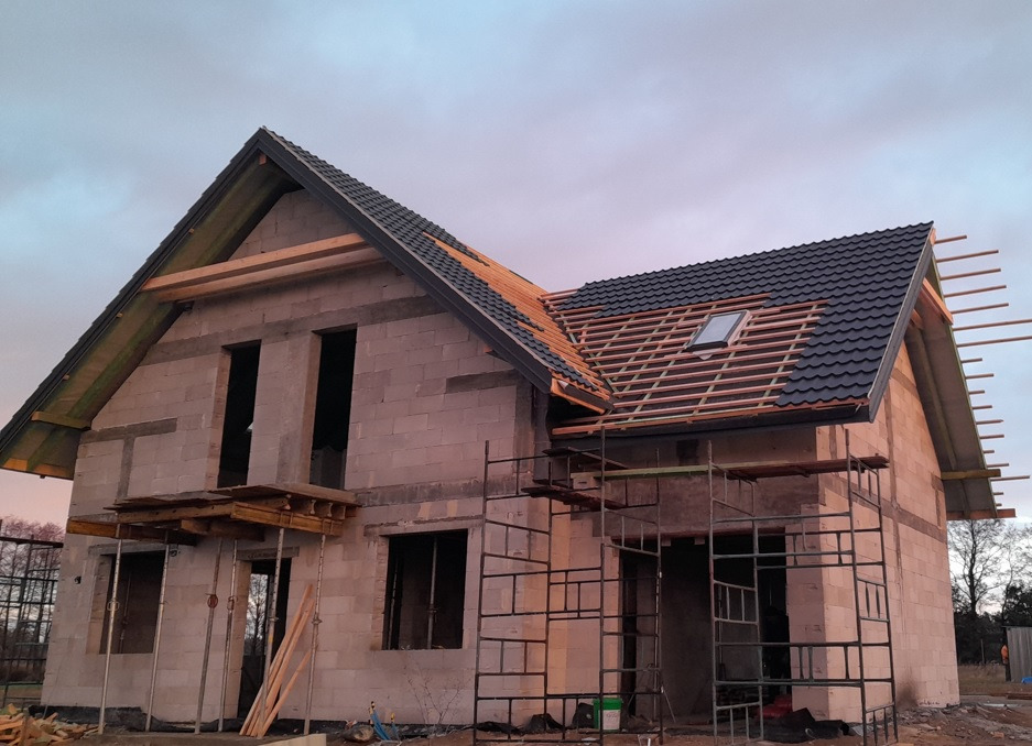Dom murowany w trakcie budowy: ściany z betonu komórkowego, częściowo pokryty czarną dachówką, widoczne rusztowania i drewniane elementy konstrukcyjne dachu.