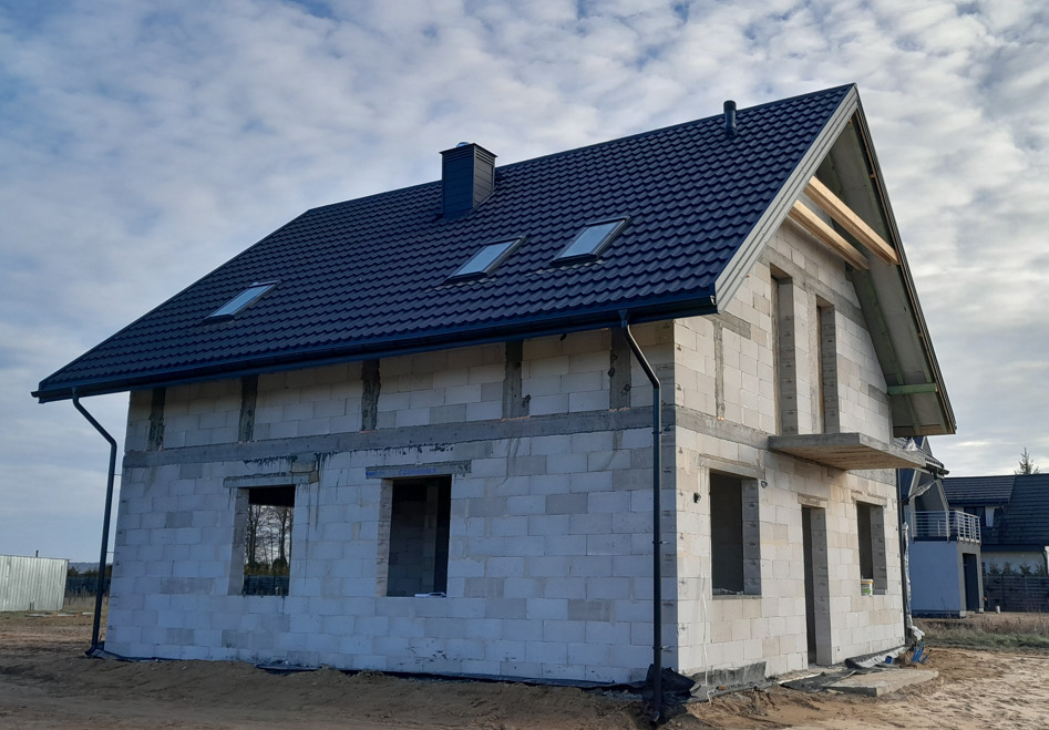 Niedokończony dom jednorodzinny z białych bloczków, z zamontowaną ciemną dachówką, oknami dachowymi i rynnami, widoczne otwarte otwory okienne i drzwiowe.