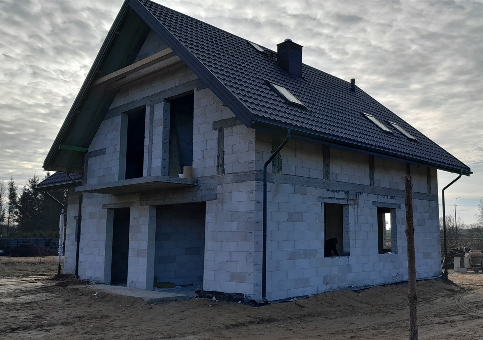 Surowy stan domu z betonu komórkowego z ciemną dachówką, widoczne otwory okienne i drzwiowe, orynnowanie i komin, na tle pochmurnego nieba.