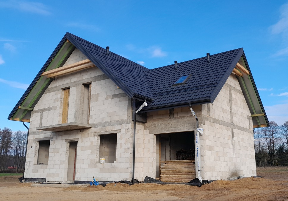 Dwukondygnacyjny budynek w stanie surowym otwartym z ciemną dachówką, widoczne niezabezpieczone rynny i drewniane elementy konstrukcji dachu.