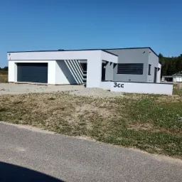 Nowoczesny, parterowy dom w kolorze białym i szarym z płaskim dachem, garażem z ciemną bramą i numerem 3cc na elewacji, otoczony niezagospodarowanym terenem.