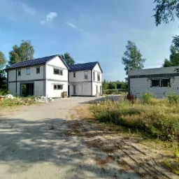 Dwa surowe, dwukondygnacyjne domy w stanie deweloperskim, z widoczną konstrukcją z pustaków i czarną dachówką, na tle zieleni i błękitnego nieba, z widocznym fragmentem drogi gruntowej.