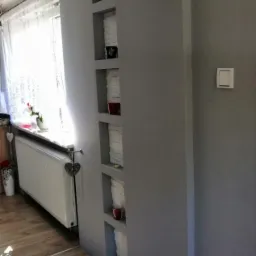 Szara ściana z wnękami dekoracyjnymi, w których umieszczono białe ręczniki i czerwone świeczki, widoczny fragment okna z firanką i grzejnik.