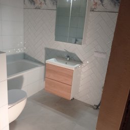 Jarosław Powroźnik - Wykończona łazienka z białą armaturą, drewnianą szafką pod umywalką, białą glazurą ułożoną w jodełkę i dekoracyjnym pasem z motywem roślinnym.