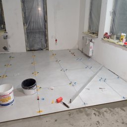 Jarosław Powroźnik - Układanie jasnych płytek podłogowych w pokoju, z użyciem poziomicy, krzyżyków dystansowych i młotka gumowego. Okna i drzwi zabezpieczone folią.