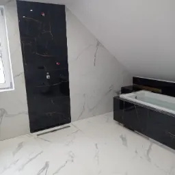 Elegancka łazienka na poddaszu z wanną obłożoną czarnym marmurem i jasnymi płytkami z marmurowym wzorem, widoczne detale instalacji.