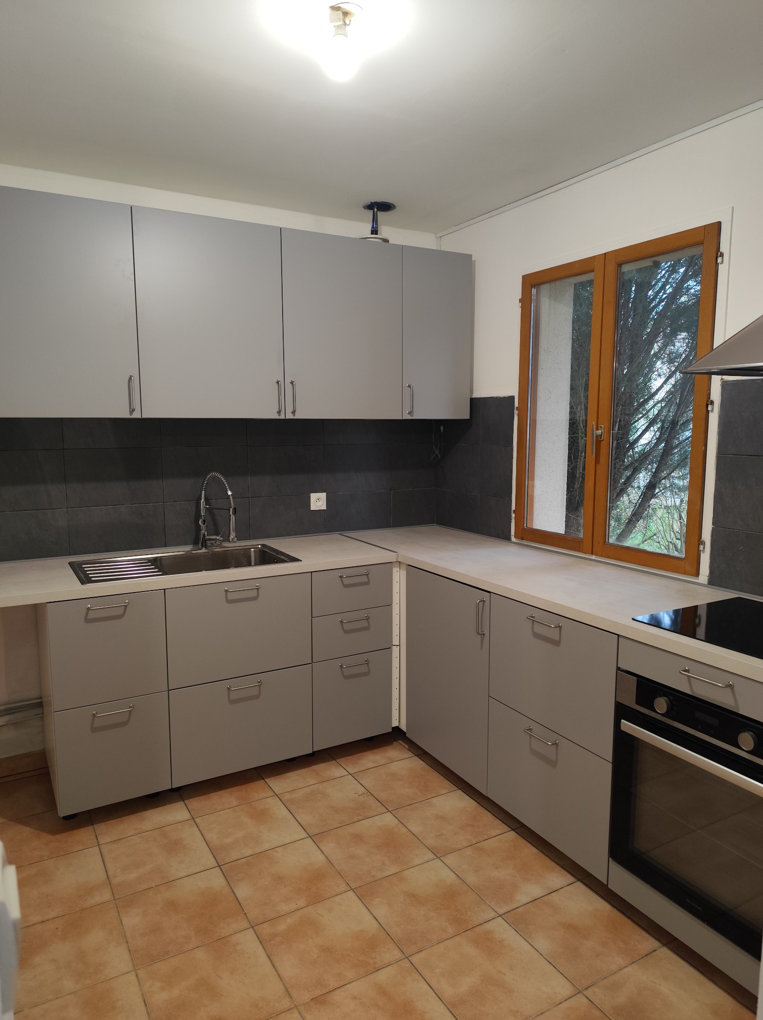 Szara, nowoczesna kuchnia z ciemnoszarą mozaiką nad blatem i brązową podłogą z terakoty. Widok na okno z drewnianą ramą i drzewo na zewnątrz.