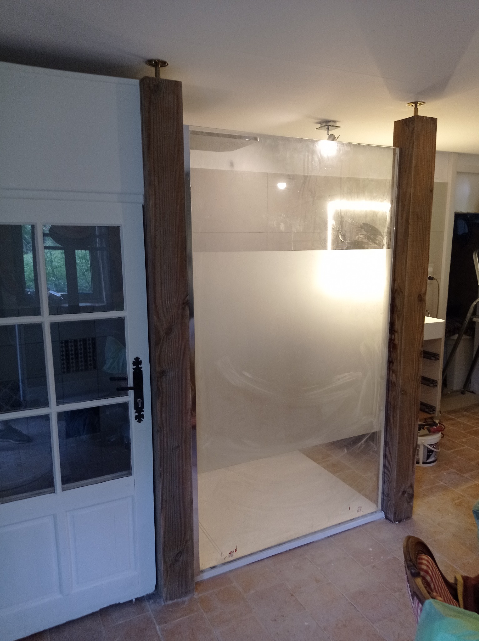 Nowoczesna kabina prysznicowa z matowego szkła, drewniane belki podtrzymujące, widoczna biała stolarka drzwiowa i terakota podłogowa w łazience.