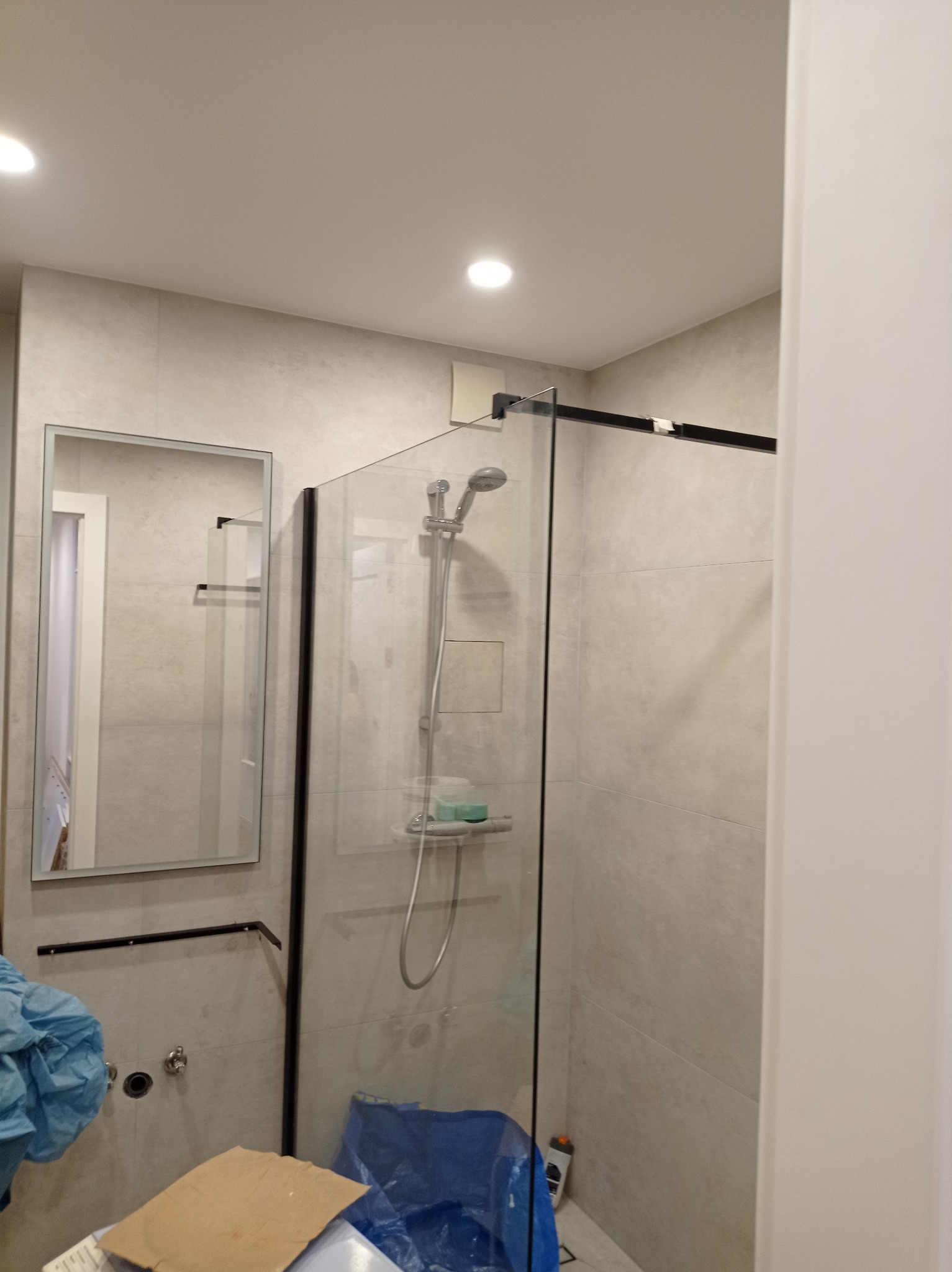 Nowoczesna kabina prysznicowa z czarnymi profilami i lustro w wykończonej łazience. Częściowo widoczne materiały budowlane. Świeże, jasne wnętrze.