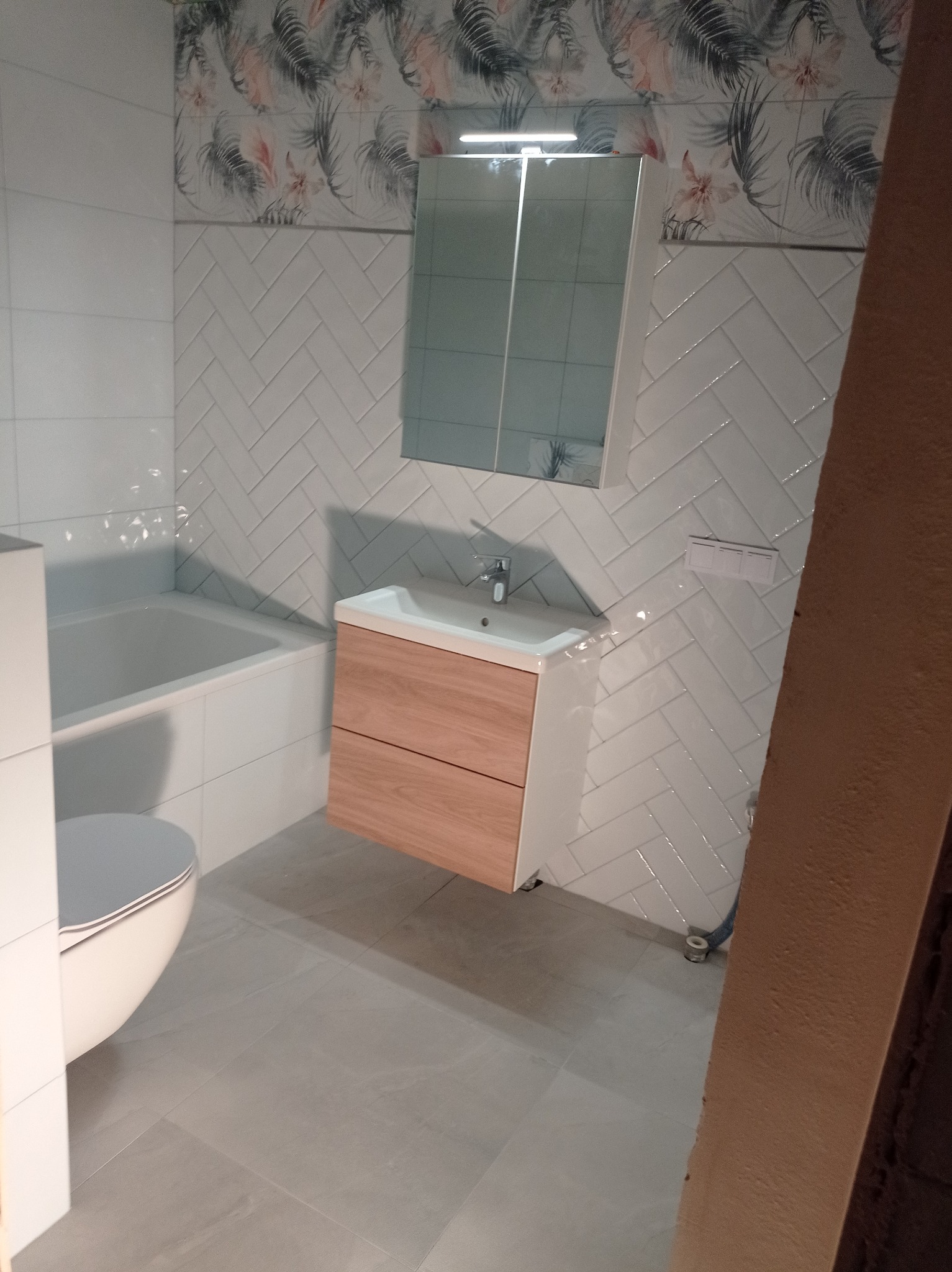 Wykończona łazienka z białą armaturą, drewnianą szafką pod umywalką, białą glazurą ułożoną w jodełkę i dekoracyjnym pasem z motywem roślinnym.