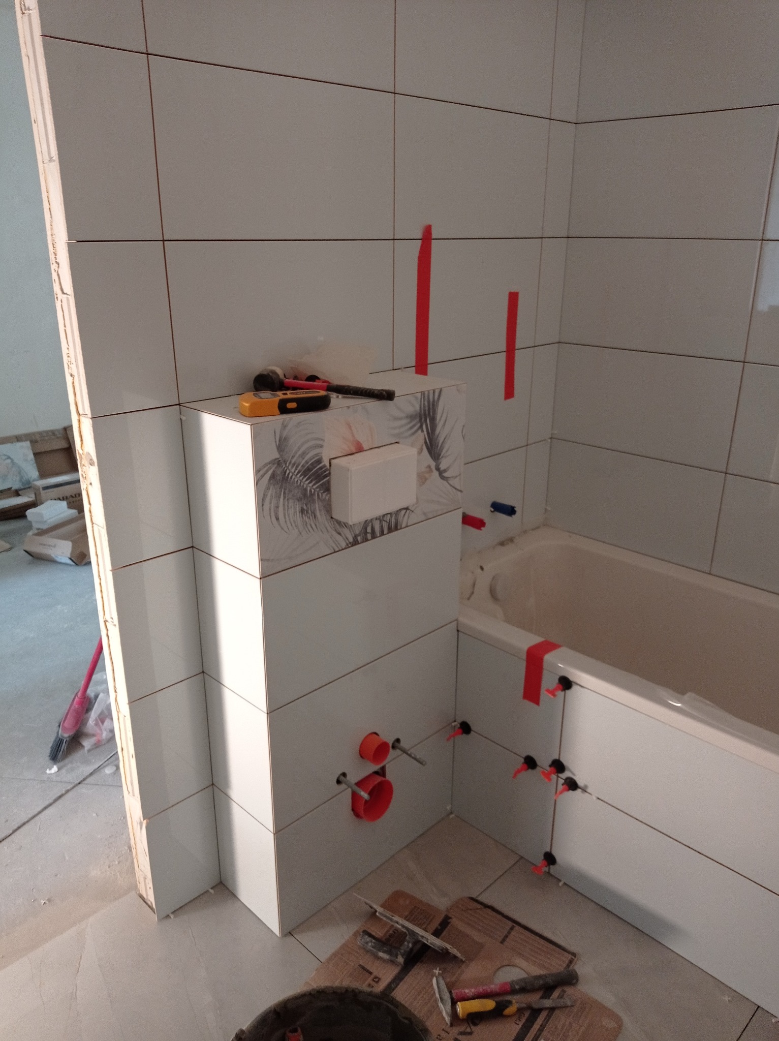 Białe płytki w łazience w trakcie montażu, widoczne narzędzia glazurnicze, poziomica, czerwone kliny i taśmy. Częściowo wykończona obudowa wanny.