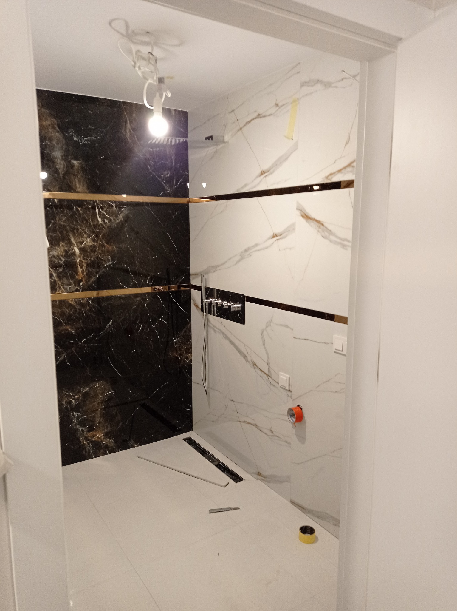 Wykończona łazienka z czarno-białymi płytkami imitującymi marmur, złotymi akcentami i odpływem liniowym w podłodze. Widoczne narzędzia glazurnicze.