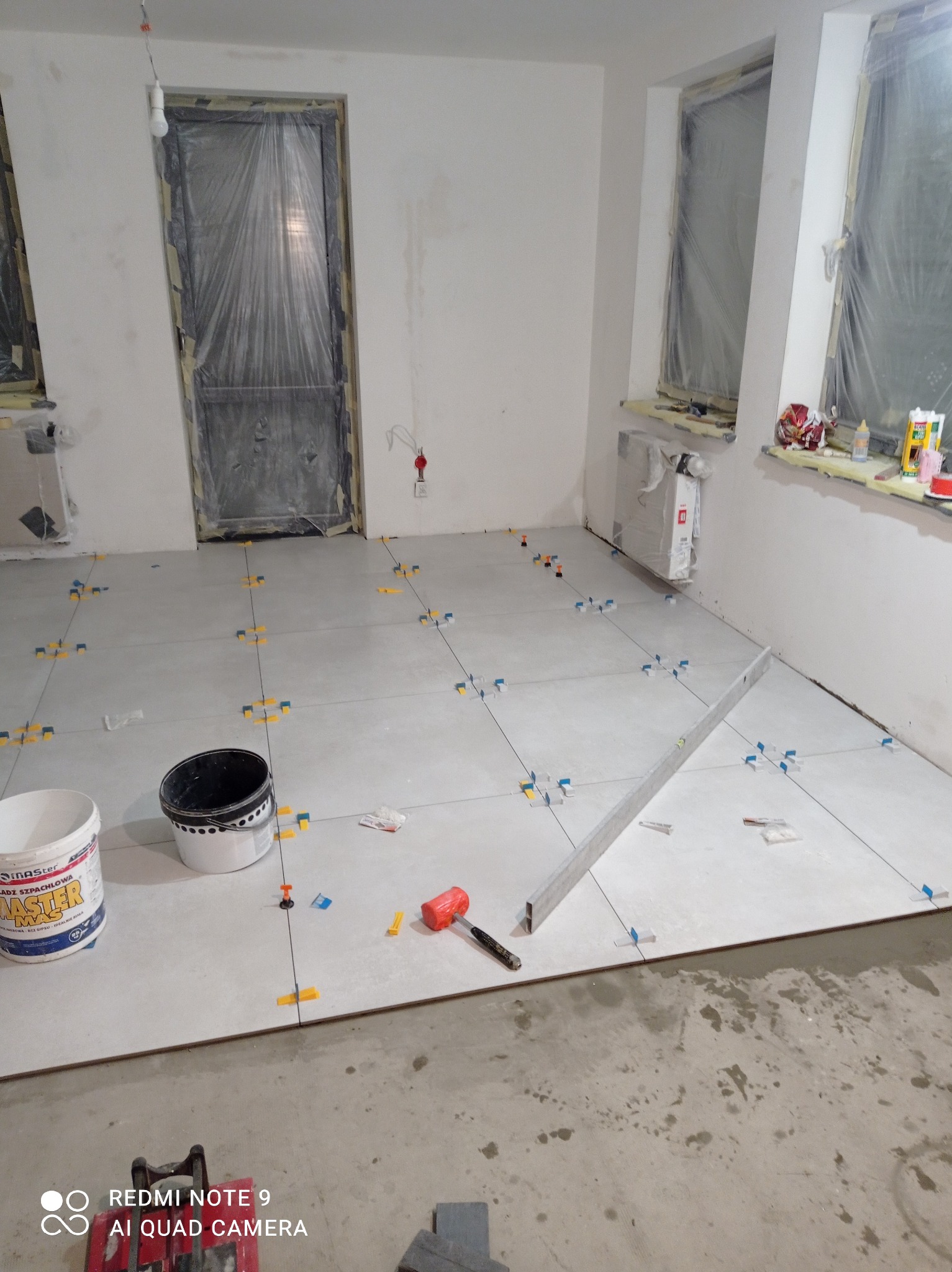 Układanie jasnych płytek podłogowych w pokoju, z użyciem poziomicy, krzyżyków dystansowych i młotka gumowego. Okna i drzwi zabezpieczone folią.
