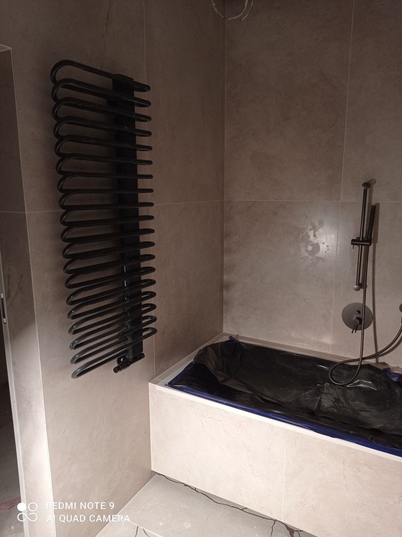 Nowoczesna łazienka w trakcie wykończenia: czarny grzejnik drabinkowy, beżowe płytki marmurowe, bateria prysznicowa w kolorze czarnym, wanna zabezpieczona folią.