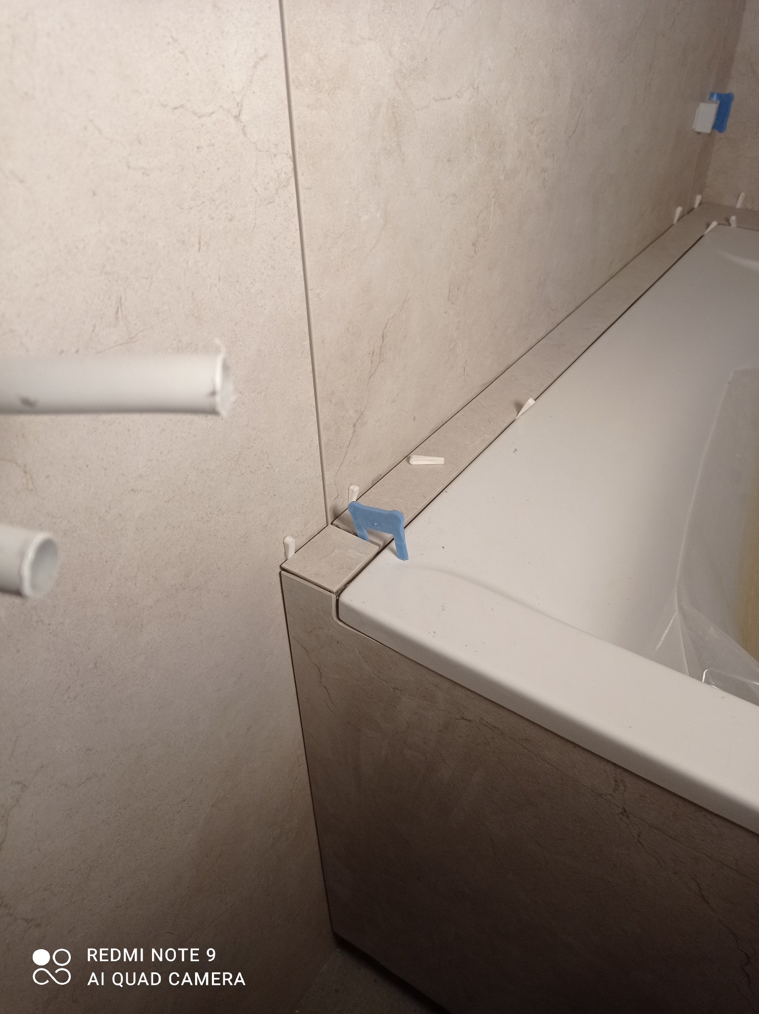 Wykończenie obudowy wanny beżowymi płytkami, z użyciem krzyżyków dystansowych. Widoczne fragmenty rur i zabezpieczona wanna. Detal z wykończenia łazienki.