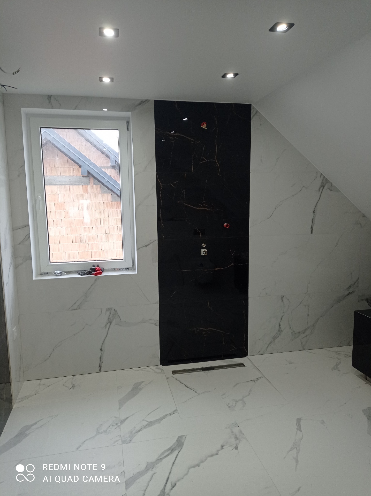 Elegancka łazienka na poddaszu z czarno-białymi płytkami imitującymi marmur. Widoczny odpływ liniowy i okno z narzędziami na parapecie.