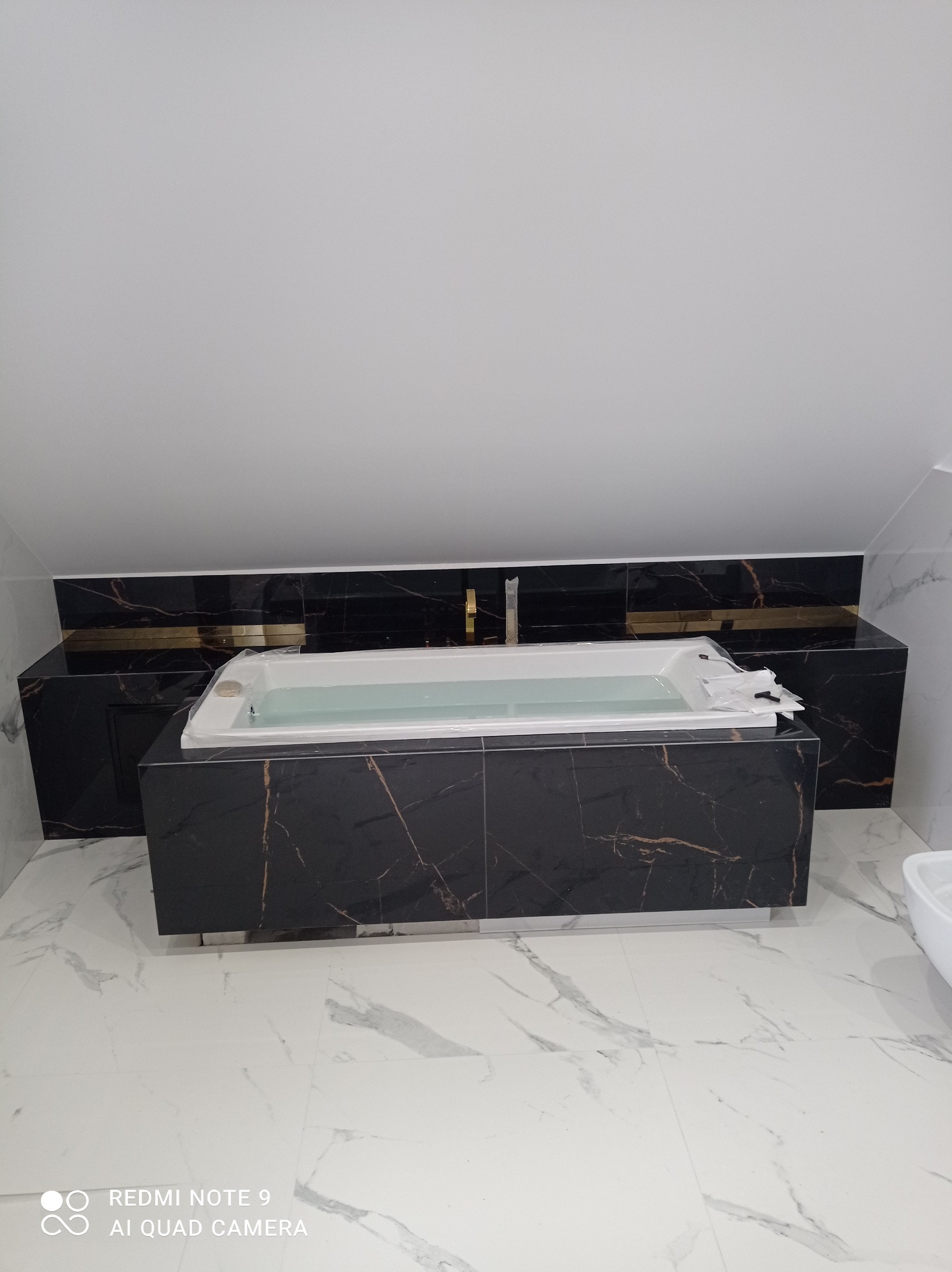 Nowoczesna łazienka na poddaszu z wanną obudowaną czarnym marmurem i białymi płytkami podłogowymi. Widoczny fragment bidetu. Ukończona aranżacja wnętrza.