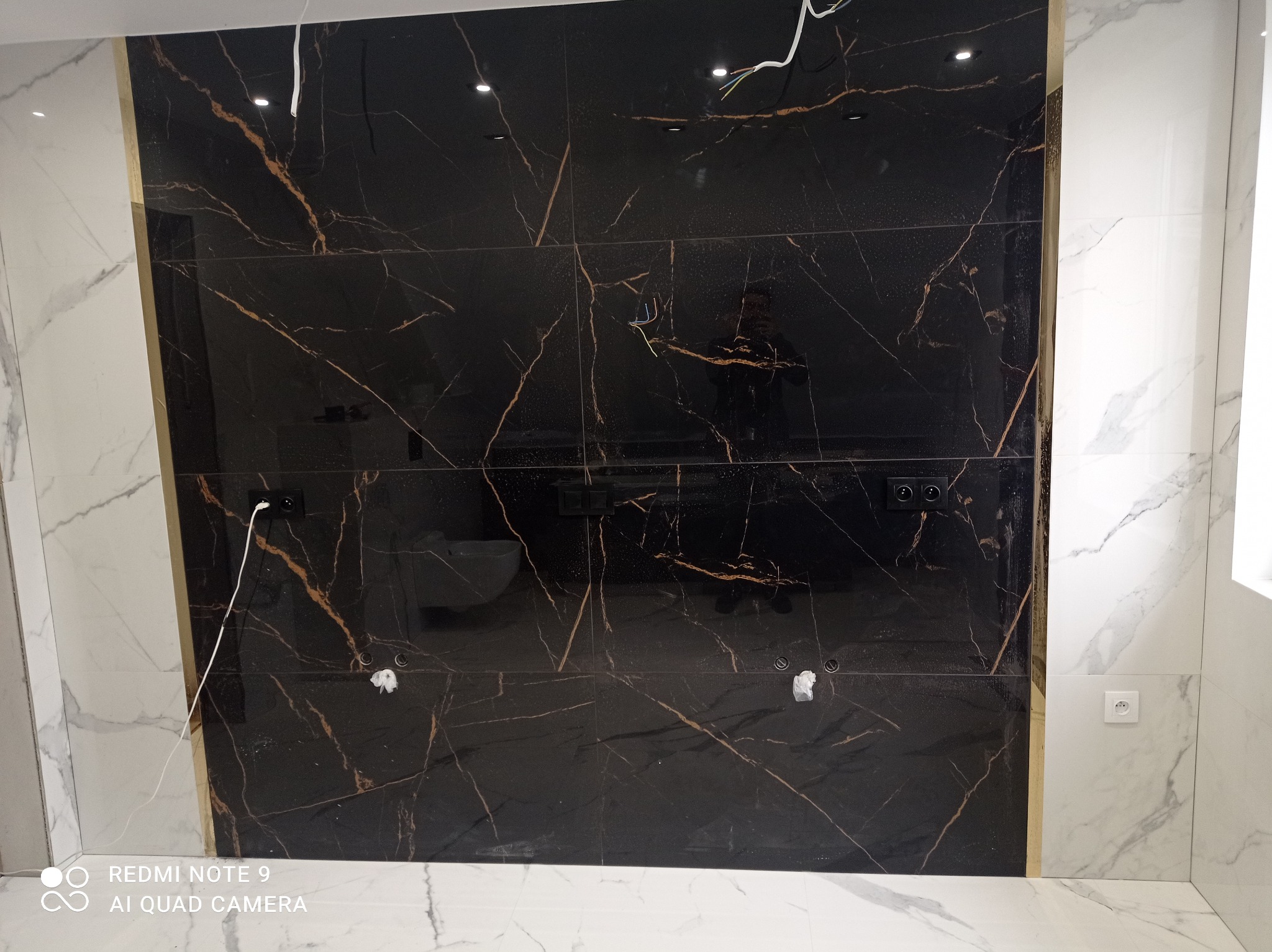 Ściana wyłożona czarnymi płytkami z brązowymi żyłami, kontrastująca z białymi marmurowymi płytkami. Widoczne gniazdka elektryczne i wyprowadzenia instalacji.