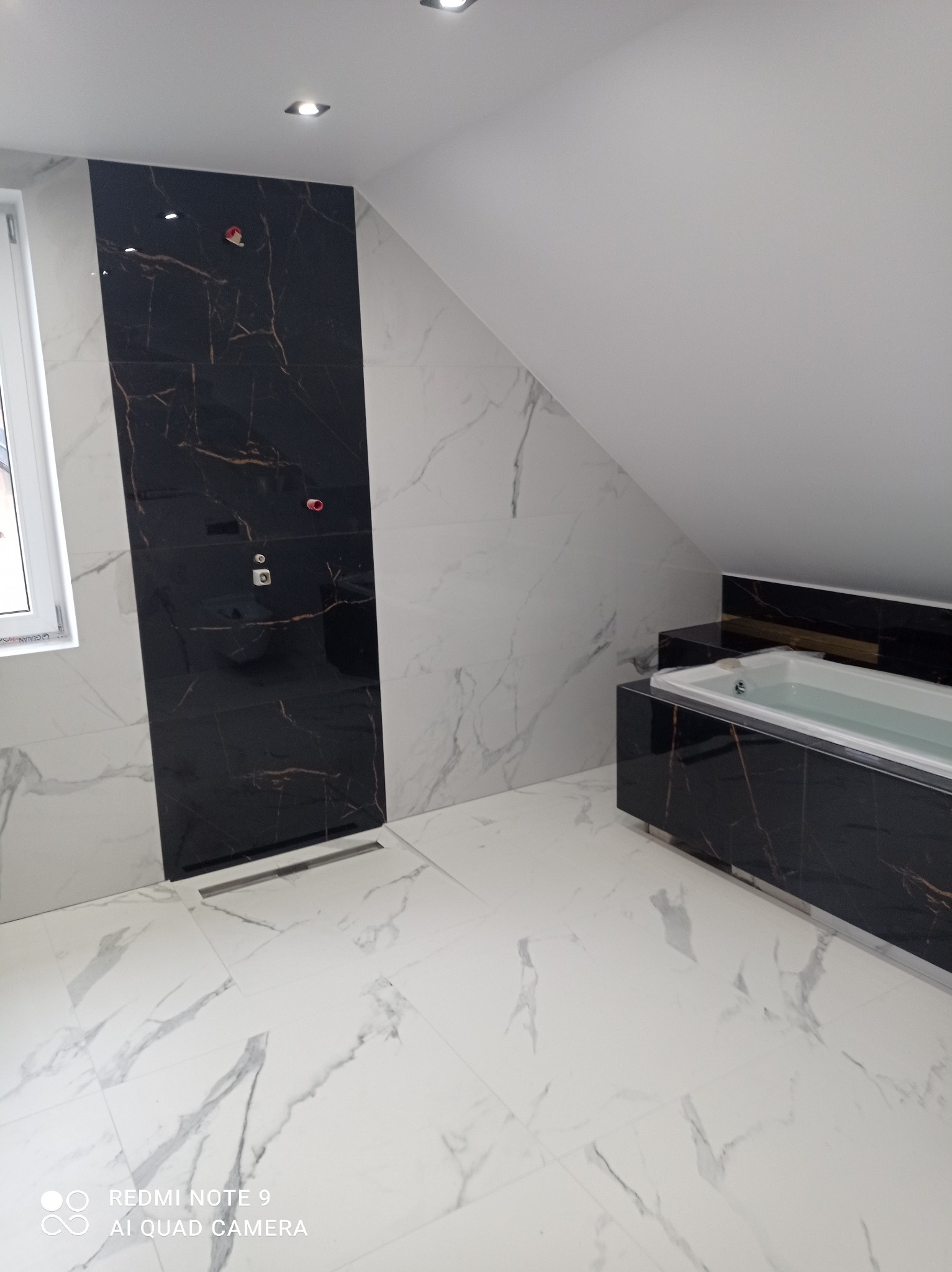 Elegancka łazienka na poddaszu z wanną obłożoną czarnym marmurem i jasnymi płytkami z marmurowym wzorem, widoczne detale instalacji.