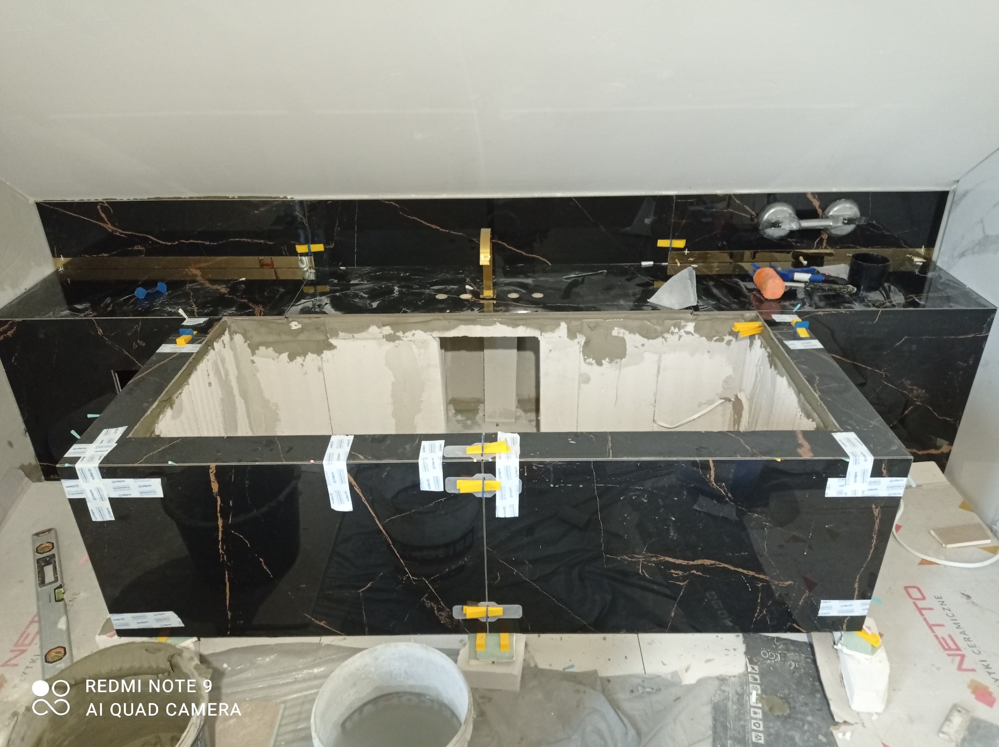 Montaż czarnych płytek imitujących marmur z złotymi żyłami na zabudowie umywalki, widoczne poziomica i narzędzia glazurnicze. Etap wykończeniowy łazienki.