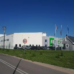 Szeroki kąt budynku przemysłowego z białą elewacją i rzędem okien, obok mniejszy budynek biurowy z elementami zieleni i trzema flagami na tle błękitnego nieba, z zaparkowanymi samochodami i znakiem...