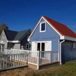 Dwa małe domki z drewnianą elewacją, jeden niebieski z czerwoną dachówką i białymi okiennicami, drugi szary z ciemną dachówką, połączone białym drewnianym ogrodzeniem.