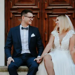 Uśmiechnięta para młoda siedzi na kamiennym murku na tle dużych, drewnianych drzwi; panna młoda w koronkowej sukni i welonie, pan młody w granatowym garniturze z muszką.