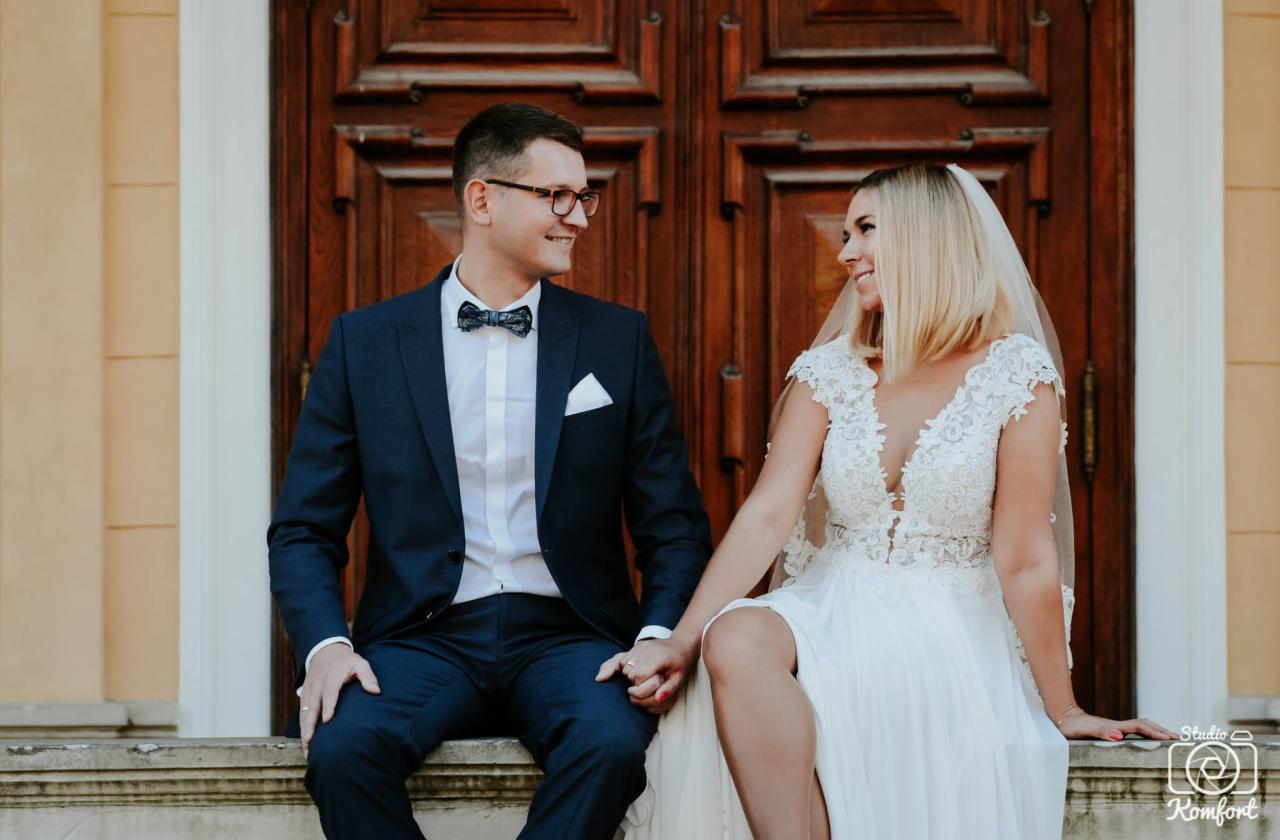 Uśmiechnięta para młoda siedzi na kamiennym murku na tle dużych, drewnianych drzwi; panna młoda w koronkowej sukni i welonie, pan młody w granatowym garniturze z muszką.