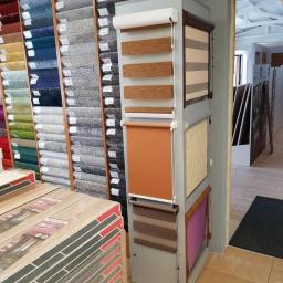 Bopan Żurawski Artur Żurawska Katarzyna S.C. - Ekspozycja rolet okiennych w sklepie z wykładzinami i panelami, obok ułożone paczki paneli podłogowych.