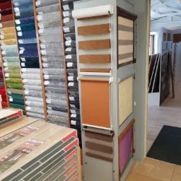 Ekspozycja rolet okiennych w sklepie z wykładzinami i panelami, obok ułożone paczki paneli podłogowych.