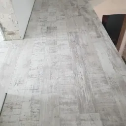 Ułożona podłoga z paneli o wzorze imitującym postarzane, bielone drewno w trakcie remontu. Widoczne surowe ściany i fragment schodów.