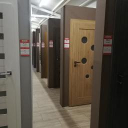Bopan Żurawski Artur Żurawska Katarzyna S.C. - Wystawa rzędu drzwi wewnętrznych o różnych kolorach i wzorach, z etykietami cenowymi, ustawionych wzdłuż korytarza z podłogą imitującą drewno.