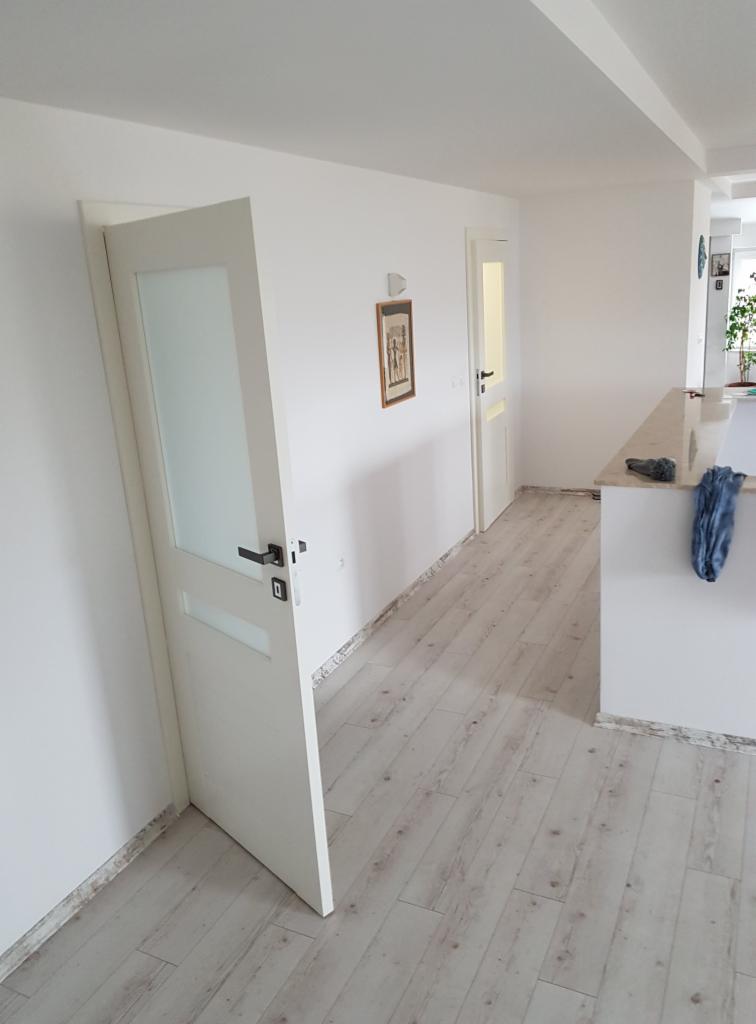 Białe drzwi wewnętrzne z matową szybą, czarna klamka, widoczne panele podłogowe w jasnym odcieniu drewna, fragment ściany z obrazkiem, wnętrze mieszkania.