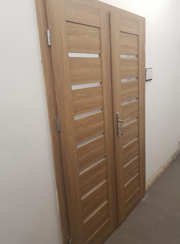 Dwa skrzydła drzwi wewnętrznych z jasnego drewna, z poziomymi, prostokątnymi przeszkleniami, ustawione obok białej ściany w trakcie montażu lub przechowywania.