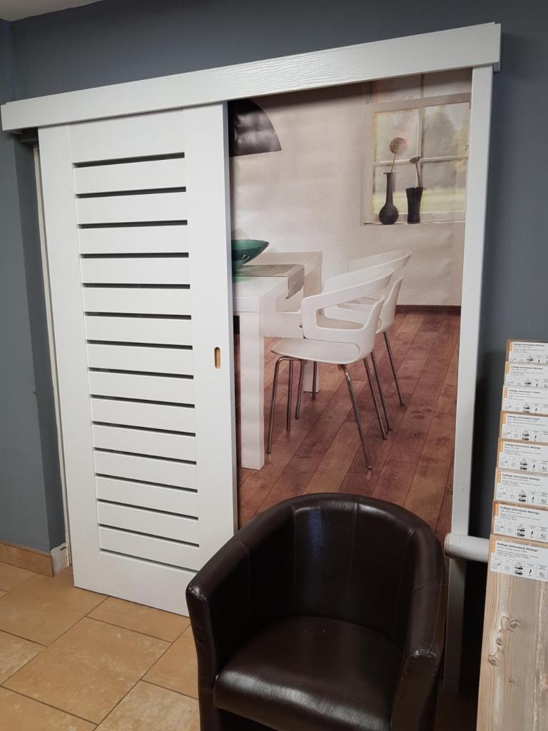 Białe drzwi przesuwne z poziomymi szczelinami, częściowo zasłaniające fototapetę przedstawiającą jadalnię z białymi krzesłami i drewnianą podłogą; w tle szara ściana i brązowy fotel.