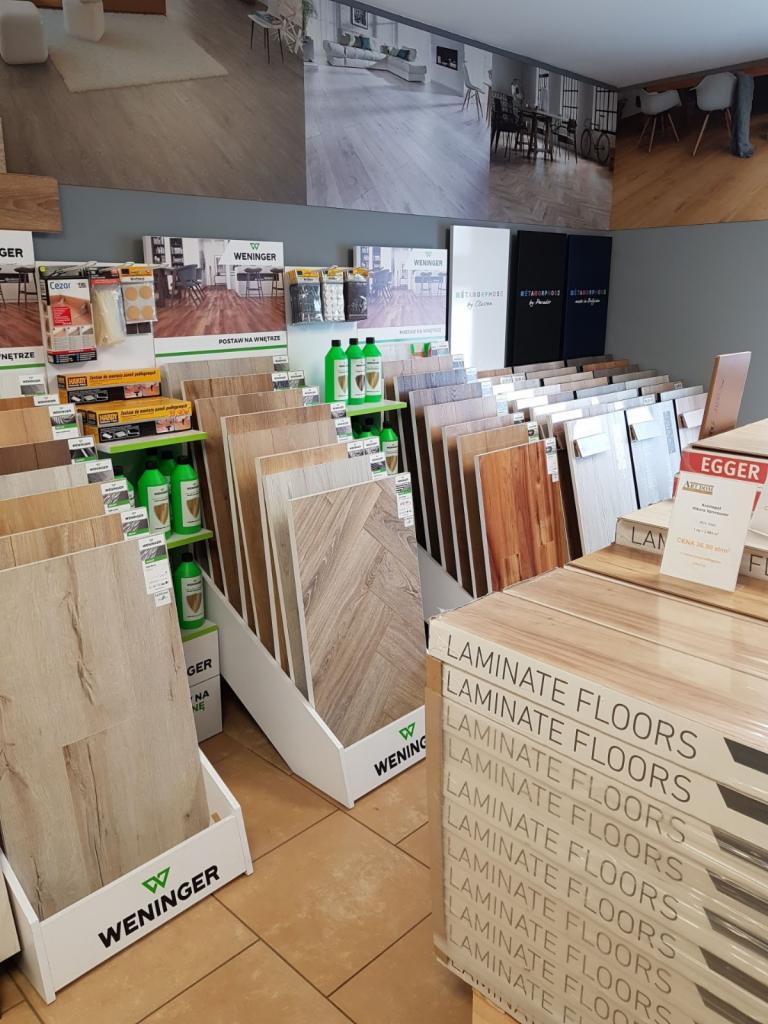 Ekspozycja paneli podłogowych w sklepie, różne wzory i kolory na stojakach marki Weninger, w tle aranżacje wnętrz z wykorzystaniem tych paneli.