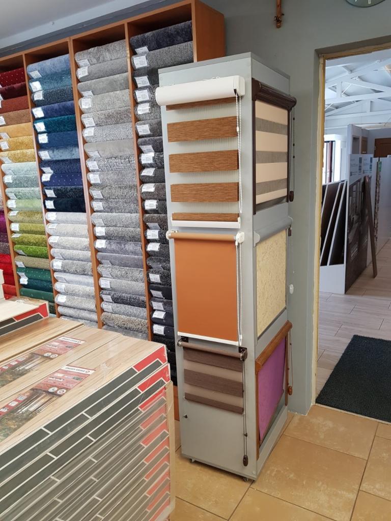 Ekspozycja rolet okiennych w sklepie z wykładzinami i panelami, obok ułożone paczki paneli podłogowych.