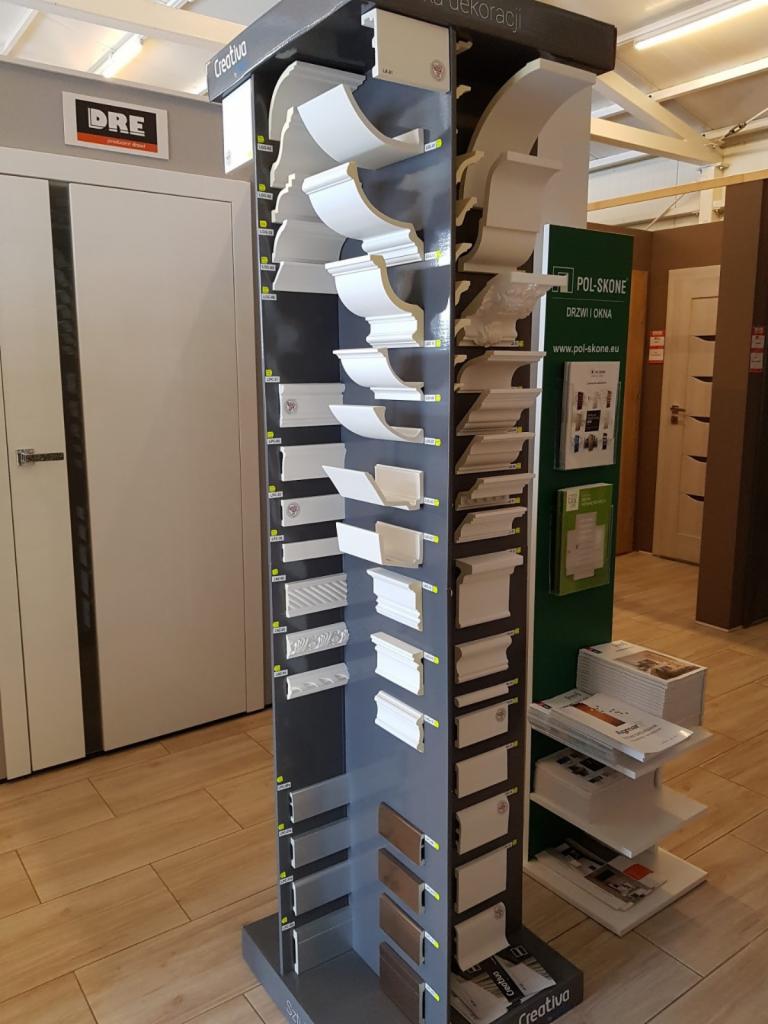 Ekspozycja różnych profili dekoracyjnych, listew przypodłogowych i wykończeniowych w sklepie, z widocznym logo producenta Creativa i drzwi w tle.