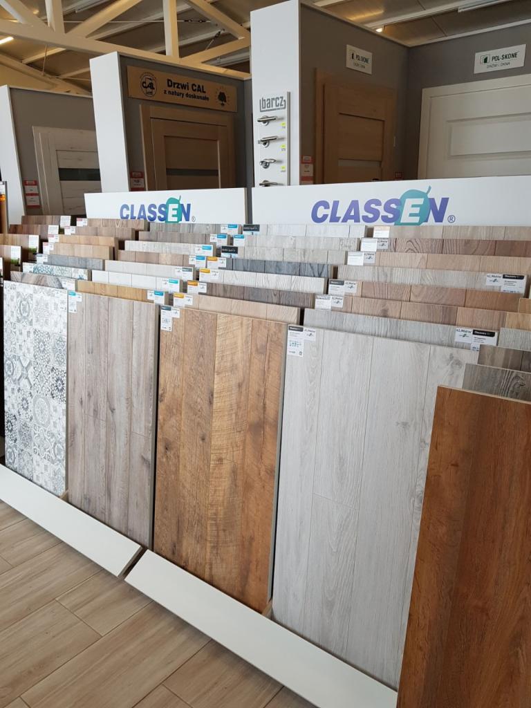 Ekspozycja próbek paneli podłogowych o różnych wzorach i kolorach w sklepie, z widocznym logo Classen na górze ekspozycji i ekspozycją drzwi w tle.
