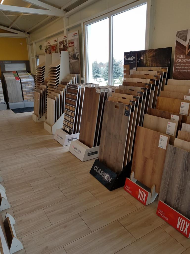 Ekspozycja różnych wzorów paneli podłogowych w sklepie, widoczne marki Classen, Vox i Swiss Krono, ułożone na podłodze wyłożonej płytkami imitującymi drewno, z oknem w tle.