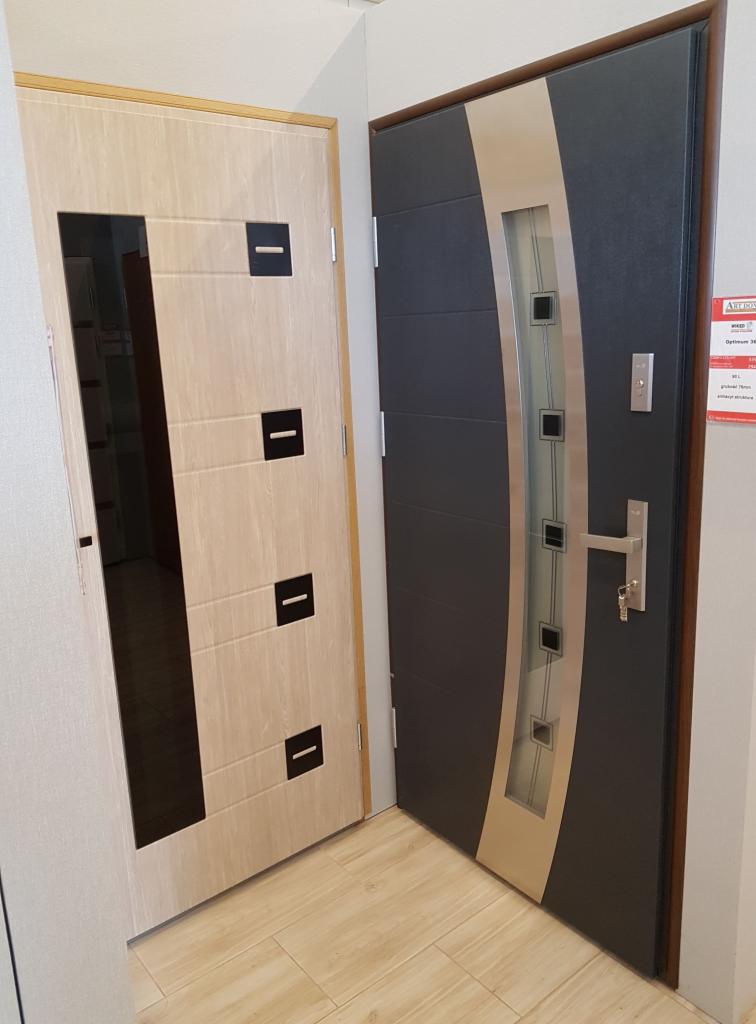 Dwa modele drzwi wewnętrznych: jasne drewnopodobne z czarnymi wstawkami i ciemne grafitowe z przeszkleniem i srebrnymi akcentami, prezentowane w salonie sprzedaży.